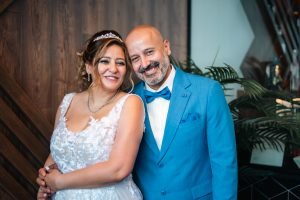 Happy newlywed couple embracing, with the bride in a white lace wedding dress and the groom in a blue suit, standing in front of a wooden wall and green plants.