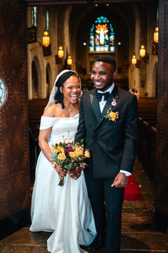 A beautiful Black wedding couple walking down the aisle in a stunning Church of St. Andrew and St. Paul, surrounded by elegant stained glass windows and warm lighting. The bride wears a gorgeous white off-the-shoulder gown with a flowing veil, holding a vibrant bouquet of yellow roses and greenery. The groom is dashing in his black tuxedo with a matching yellow boutonniere. Capture your special day with Keyris Rodriguez Photography—expert wedding photographers serving couples in Canada and beyond. Visit keyrisrodriguez.com to learn more.
