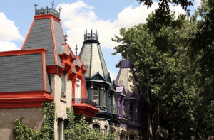 Colorful Victorian-style rooftops of historic townhouses in Le Plateau-Mont-Royal, Montreal, featuring vibrant red, teal, and purple accents under a bright summer sky, partially framed by lush green trees.